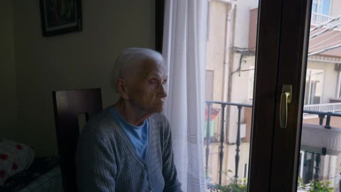 悲伤体贴的皱纹老妇人坐在阳台门的椅子上环顾四周，看着窗外。高级高加索女性退休人员在家室内思考的肖像。