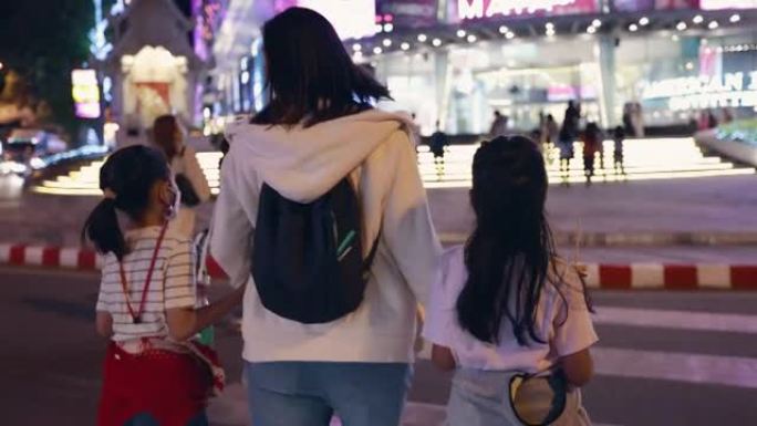 亚洲家庭的母亲和两个女儿牵手在一起，在夜间，人群和许多交通拥堵的汽车过马路。家庭在夜城玩得开心。