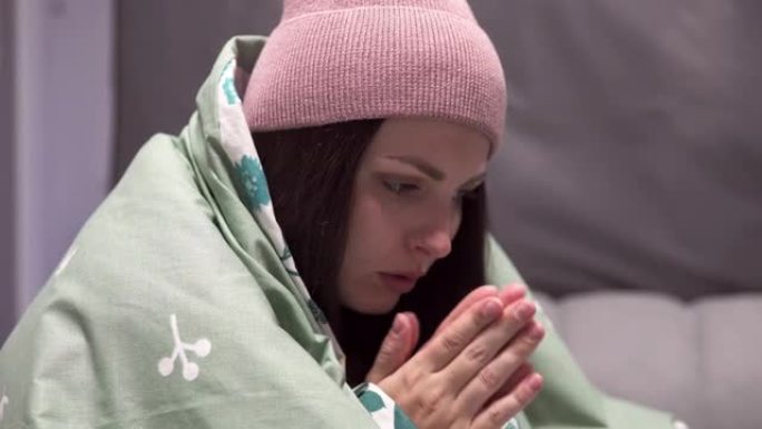 一个戴着毯子的帽子的年轻女子在一个没有暖气的公寓的床上因寒冷而颤抖。由于能源部门的危机，电力和集中供
