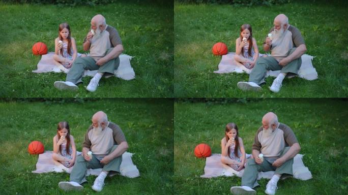 宽镜头无忧无虑的高加索祖父和孙女坐在公园绿色的夏季草地上谈论吃冰淇淋。快乐放松的高级男人和女孩在户外