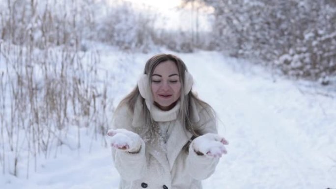 一个穿着冬衣和耳机的美丽女孩以心脏的形式从雪球的手套上吹雪。冬天在森林里放纵雪