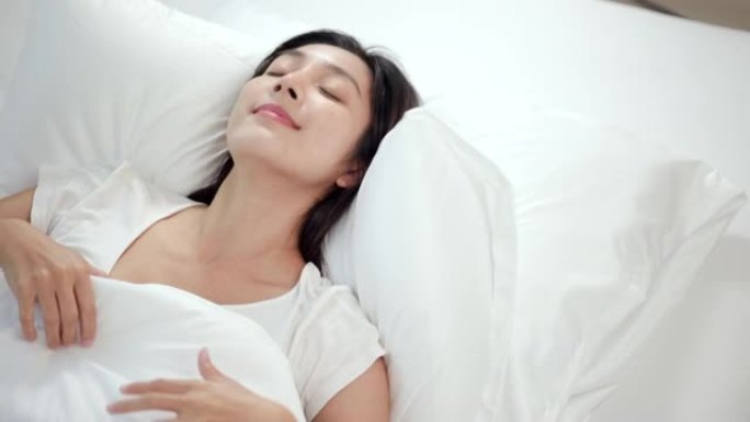 美丽的亚洲女人躺在床上。年轻女子在柔软的白色亚麻枕头上舒适舒适的新鲜床上睡得很好。早上在卧室里。健康