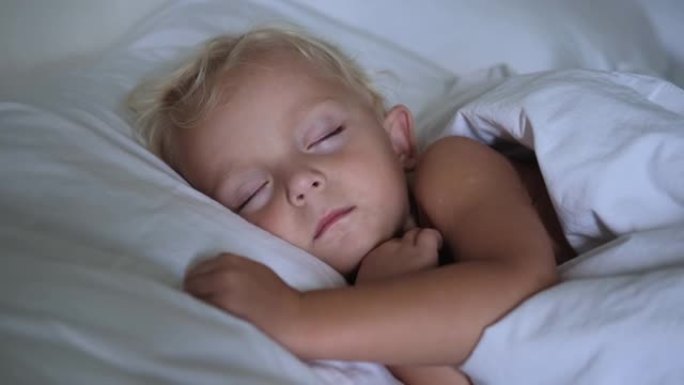 一个两岁的金发女孩安稳地睡在雪白枕头上的温暖床上，上面盖着雪白的毯子。一个小女孩做了愉快的梦。健康的