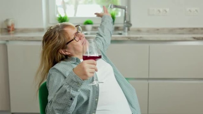 厨房里端着一杯红酒的快乐中年妇女坐在绿色的扶手椅上。一个女人在厨房的椅子上旋转。