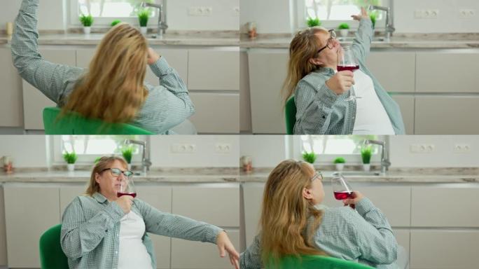 厨房里端着一杯红酒的快乐中年妇女坐在绿色的扶手椅上。一个女人在厨房的椅子上旋转。