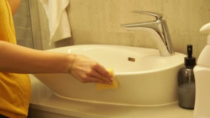 女性用海绵擦洗白色现代浴室水槽，相机变焦。女家庭主妇在洗手间打扫脏家具。用清洁剂擦拭水槽和水龙头。清