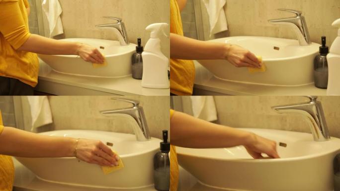 女性用海绵擦洗白色现代浴室水槽，相机变焦。女家庭主妇在洗手间打扫脏家具。用清洁剂擦拭水槽和水龙头。清