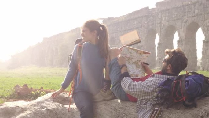 快乐的年轻夫妇背包客游客坐在原木后备箱上阅读地图指南指向罗马帕尔科德格里阿奎多蒂公园的古罗马渡槽遗址