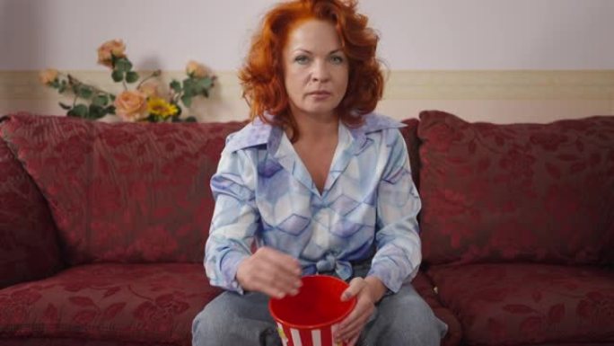 愤怒不满的复古女人扔爆米花坐在家里的沙发上看着相机。愤怒的高加索红发家庭主妇在20世纪80年代20世