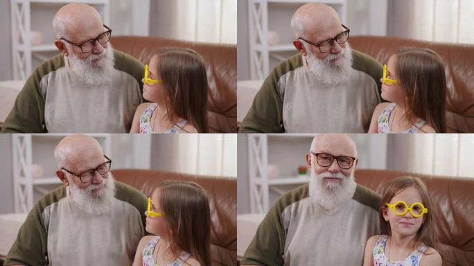 戴眼镜的高级祖父和戴黄色玩具眼镜的迷人孙女互相看着对方转身看着相机微笑。积极的高加索老人和女孩在慢动
