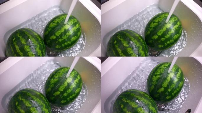 两个条纹的圆形西瓜在白色水槽下的水流，在厨房里用水和可溶性苏打水清洗成熟西瓜的俯视镜头，去除化学杂质