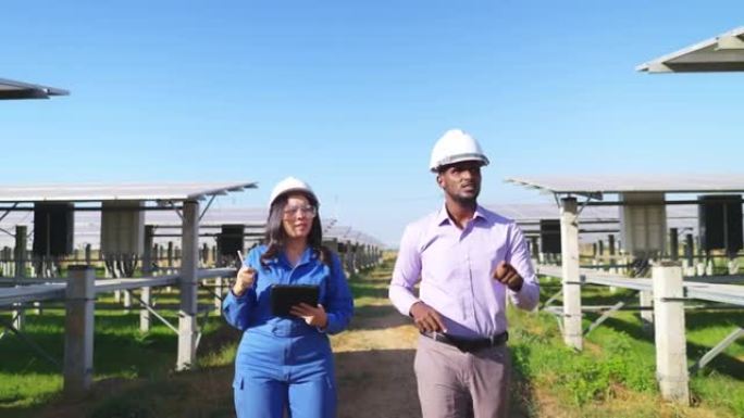 穿着蓝色防护服，安全眼镜，头盔的亚洲中年工程师在平板电脑上与非洲投资者一起在太阳能农场散步时注意