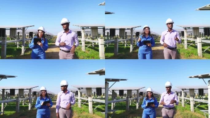 穿着蓝色防护服，安全眼镜，头盔的亚洲中年工程师在平板电脑上与非洲投资者一起在太阳能农场散步时注意