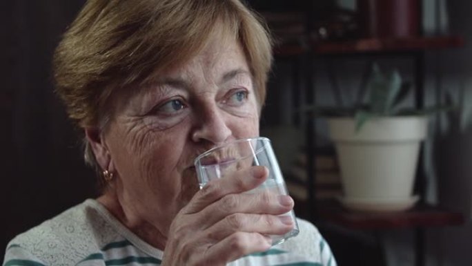 一位老年妇女在家庭环境的背景下从玻璃杯中喝水。恢复体内液体平衡