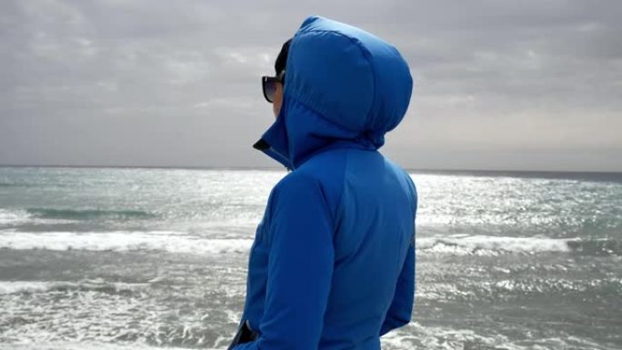 穿着蓝色夹克的孤独女人站在海边看海浪