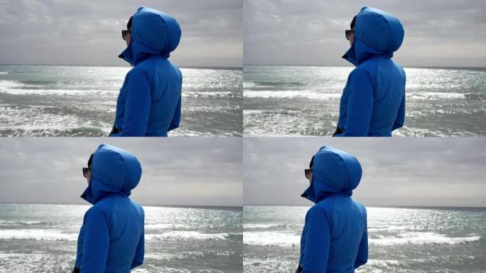 穿着蓝色夹克的孤独女人站在海边看海浪
