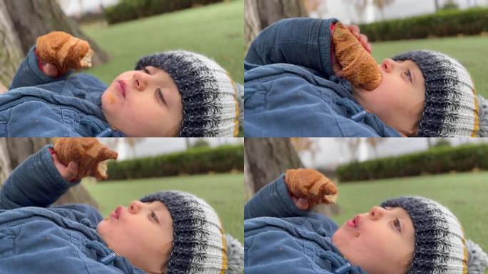 快乐的孩子在公园外面吃羊角面包。小男孩穿着冬装夹克和豆豆。儿童零食碳水化合物食品