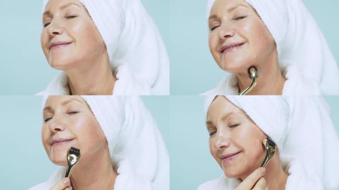 穿着白色浴袍和头上毛巾的高级女性做水疗护肤程序，使用美容滚筒在蓝色背景上进行面部按摩。美丽和自我护理