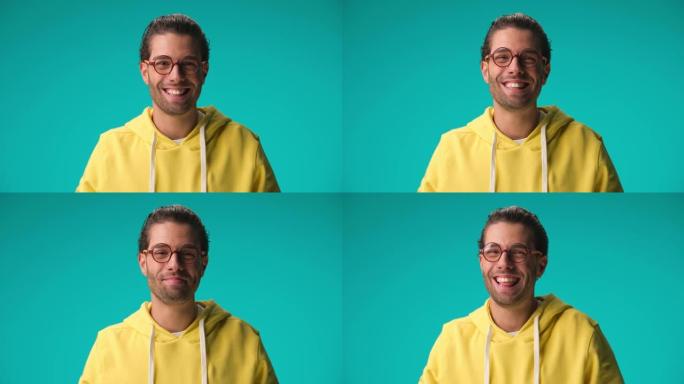 穿着黄色运动衫的害羞的年轻休闲书呆子男人戴着眼镜在蓝色背景上微笑、大笑和快乐
