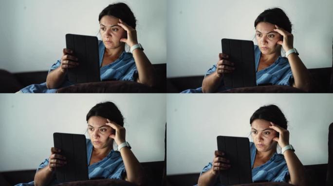 严肃的女人坐在家里的沙发上阅读设备上的信息。愤怒的人带着不满的情绪拿着平板电脑