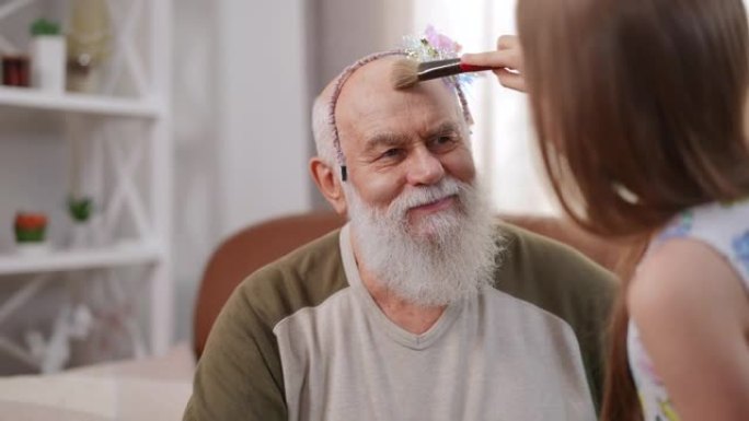 笑笑的高加索老祖父的肖像在发箍上欣赏孙女在额头上涂抹面粉。快乐放松的高级男人和迷人的女孩在室内享受周