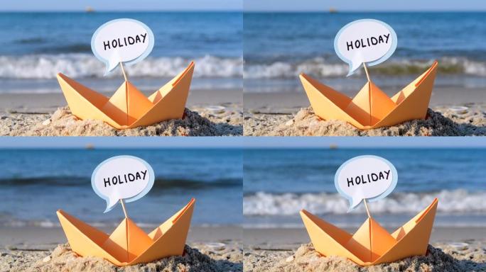 沙滩上的橙色纸船特写。阳光明媚的夏日，蓝色大海背景上的小船。坚持将单词假期插入船上。概念旅游、度假、