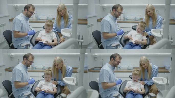 男牙医向男孩和他的母亲展示了如何以嬉戏的方式正确刷牙的下巴模型。孩子坐在牙科椅上拍手。慢动作