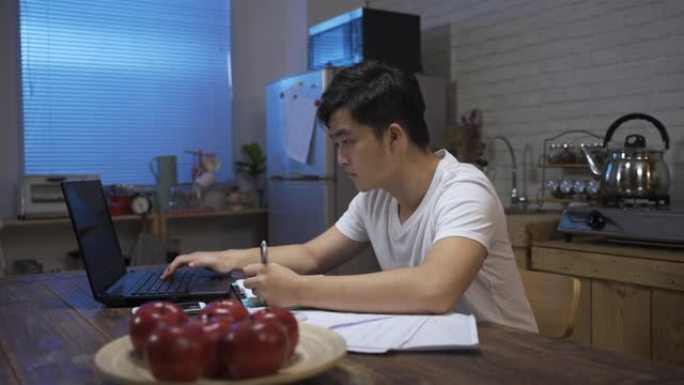 穿着休闲服在家工作到很晚的亚洲男性正在在家饭厅的计算机上做账时写笔记。