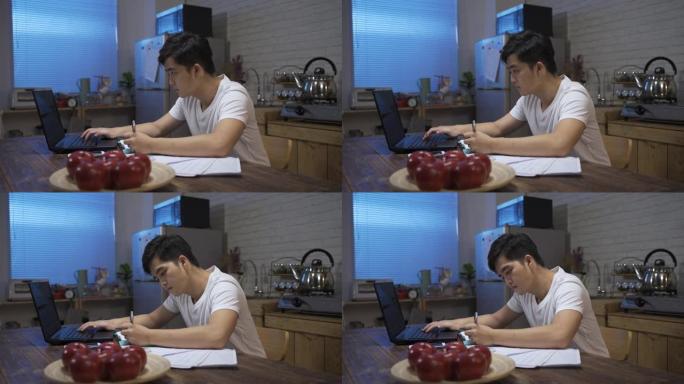 穿着休闲服在家工作到很晚的亚洲男性正在在家饭厅的计算机上做账时写笔记。
