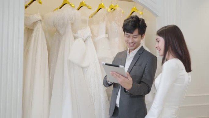 亚洲销售男士在一家服装和婚纱店使用平板电脑打开和预览婚纱，供新娘在婚纱店选择
