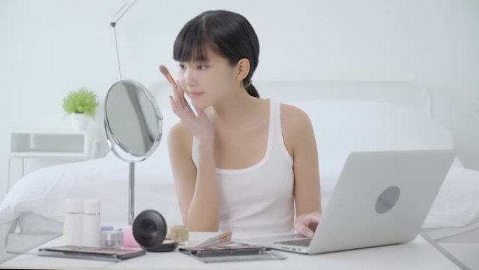年轻的亚洲女性之美，在笔记本电脑上学习用刷子在脸颊上化妆，在线辅导课程，女性教学，并在流媒体社交媒体
