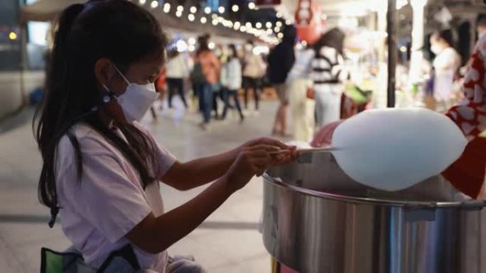 亚洲女童在街头夜市的专用机器上制作棉花糖。Kid将棉花糖棒放在机器中，将细长线形式的糖缠绕在棒上。