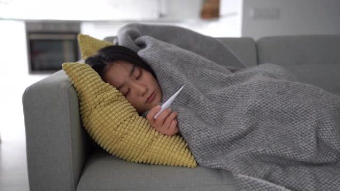 患有体温计的亚洲妇女躺在毯子下的沙发上，打喷嚏，患有流感