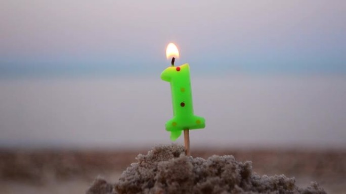 蜡烛一号在棍子上插入沙子和燃烧。模糊海浪背景下的沙子点燃蜡烛。模糊的蓝色棕色背景上的绿色燃烧蜡烛。生