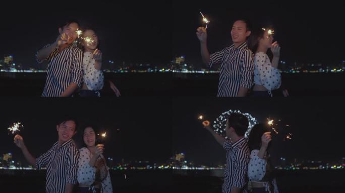 这对幸福的夫妇手持烟火灯，晚上在海滩上享受烟花汇演。