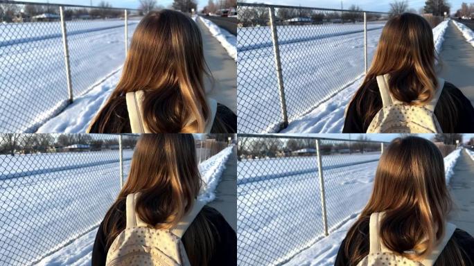 年轻女孩在校园旁边的人行道上行走的后视图，链条围栏和地面积雪