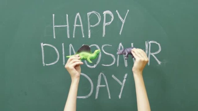 女人用粉笔写字再见学校。黑板上的恐龙日快乐，女孩的手在玩具面前玩耍