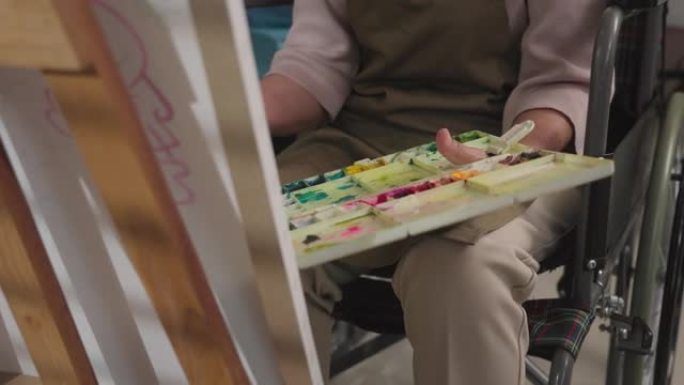 老妇人手握画笔，坐在轮椅上画画，在画布上作画