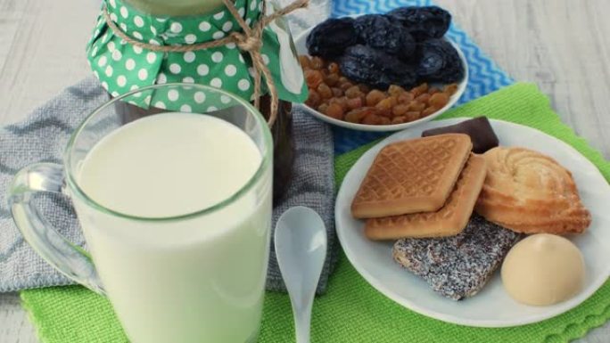 玻璃杯中的新鲜天然牛奶，一罐果酱，一个白色茶匙，一个装有干果的碟子和一个装有饼干和糖果的碟子，放在彩