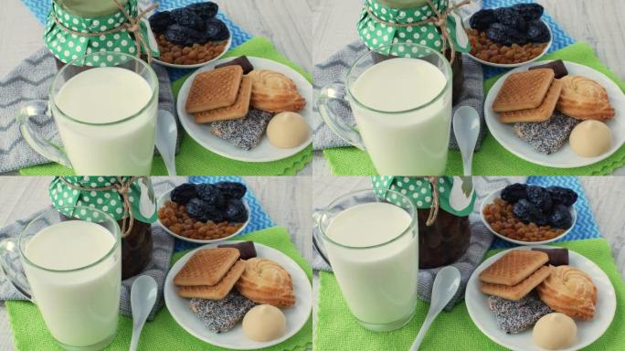 玻璃杯中的新鲜天然牛奶，一罐果酱，一个白色茶匙，一个装有干果的碟子和一个装有饼干和糖果的碟子，放在彩