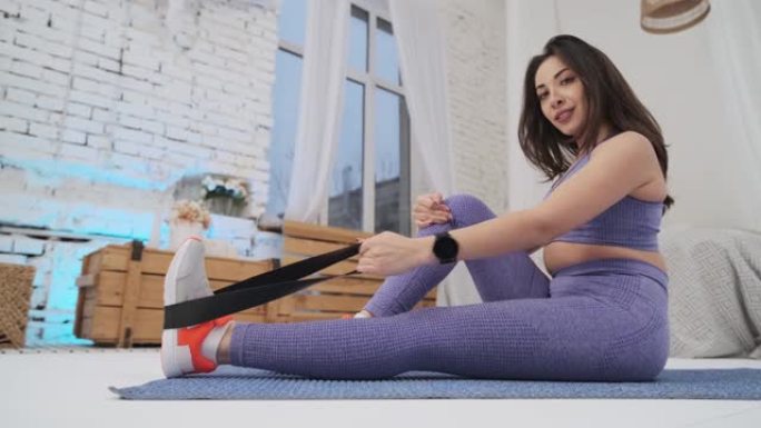 穿着紫色运动服的漂亮女孩正在家里健身。健身女孩用黑色橡皮筋在手部肌肉上锻炼。一个健康的女孩过着积极的