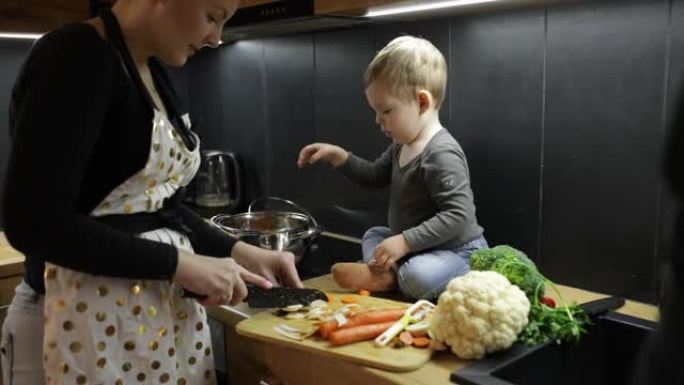 年轻女子，小婴儿和男人的家庭在家里的厨房里煮汤。母亲在桌子上切白萝卜，儿子把胡萝卜扔进锅里，父亲加欧