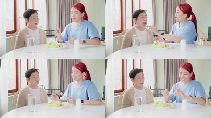穿着医疗磨砂膏的亚洲女性护理人员通过喂养素食沙拉在家吃饭来照顾亚洲老年女性。护理人员参观家庭保健概念