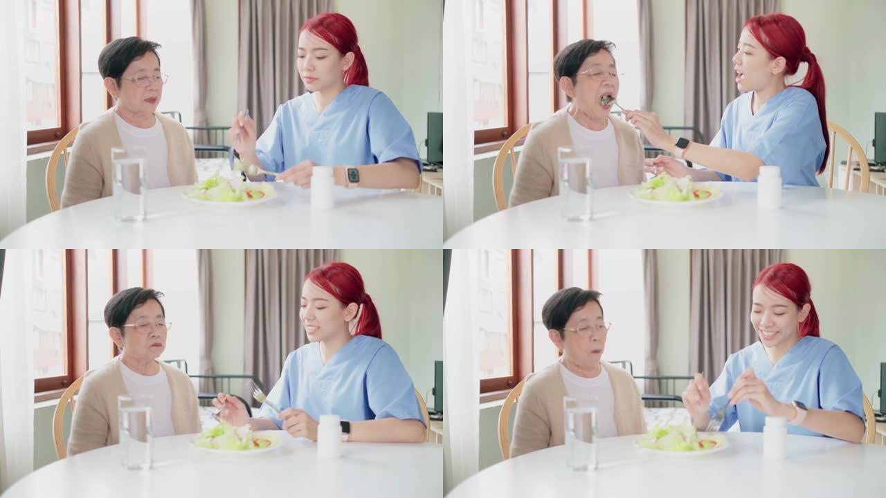 穿着医疗磨砂膏的亚洲女性护理人员通过喂养素食沙拉在家吃饭来照顾亚洲老年女性。护理人员参观家庭保健概念