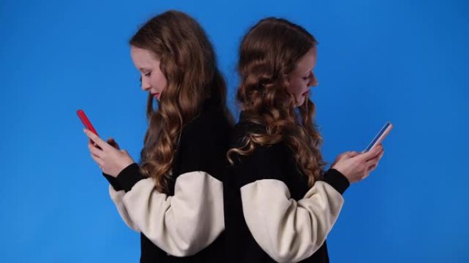 双胞胎女孩在蓝色背景下使用手机的4k视频。