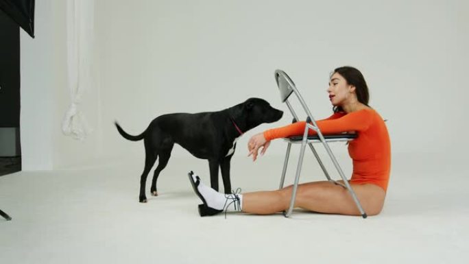 一个穿着橙色紧身衣的女孩坐在椅子上，笑着看着一只黑狗。
