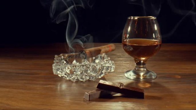 雪茄在一杯威士忌和巧克力棒附近的烟灰缸上慢慢燃烧