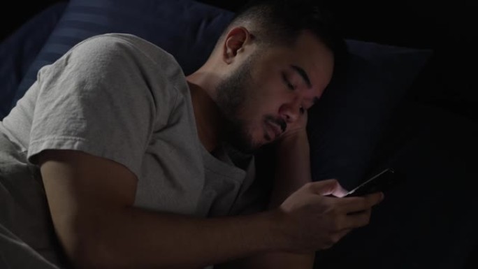 年轻的亚洲男子晚上躺在床上使用智能手机时眼睛酸痛疲惫。