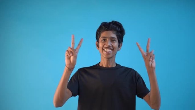 一个穿着黑色t恤的印度男孩在蓝色背景下带着灿烂的笑容享受胜利