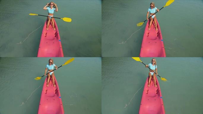 穿着时尚泳衣的漂亮运动型金发女士在异国情调的度假胜地沿着蔚蓝的海洋湾航行在粉色塑料独木舟上